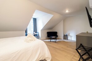 A modern bedroom with a sloped ceiling, a bed with a white duvet, a small round table with a book, a black chair, and a desk with a lamp. - Thena Hotel - Spacious Studio (Philadelphia)