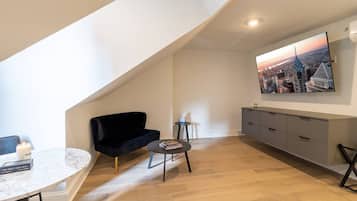 Um apartamento loft moderno com teto inclinado, sofá de veludo preto, mesa redonda pequena, mesa de centro de mármore e um quadro com vista para a cidade na parede.