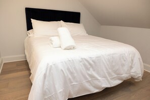 A neatly made bed with white linens and a rolled towel, set against a plain wall and wooden flooring. - Thena Hotel - Spacious Studio (Philadelphia)