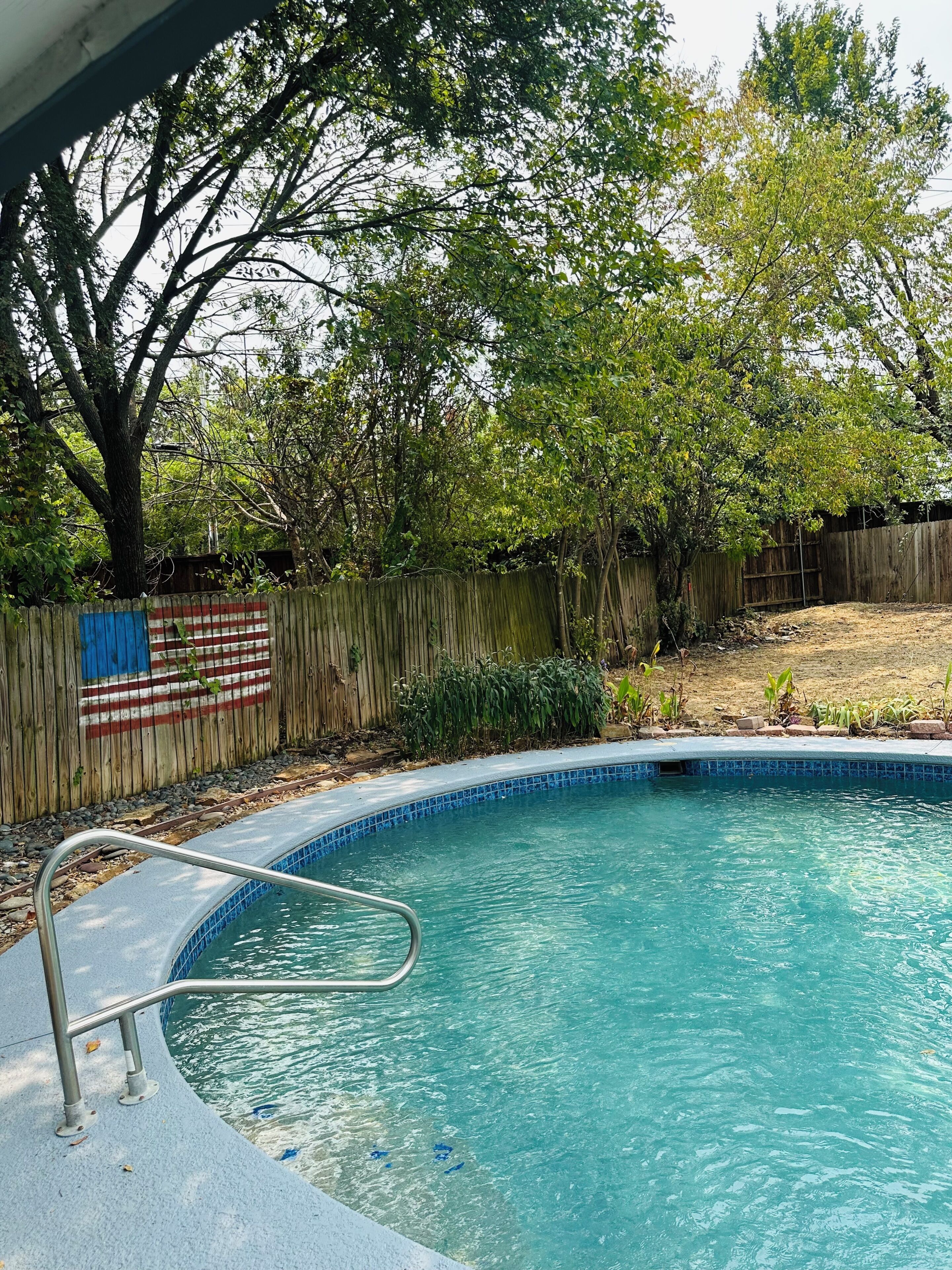 A swimming pool with a metal ladder, surrounded by a wooden fence with a flag, trees, and a grassy yard.