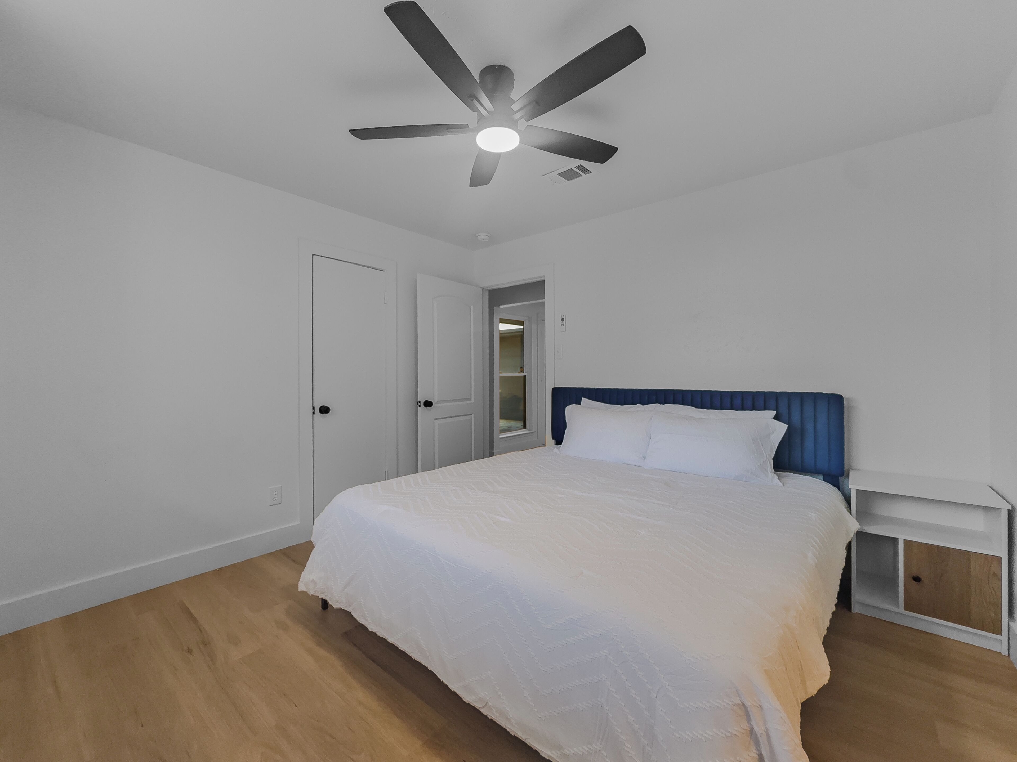 A bedroom with a bed, a nightstand, a ceiling fan, and a door.