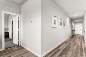 Un pasillo con piso de madera, paredes blancas y una puerta que da a una habitación con cama.