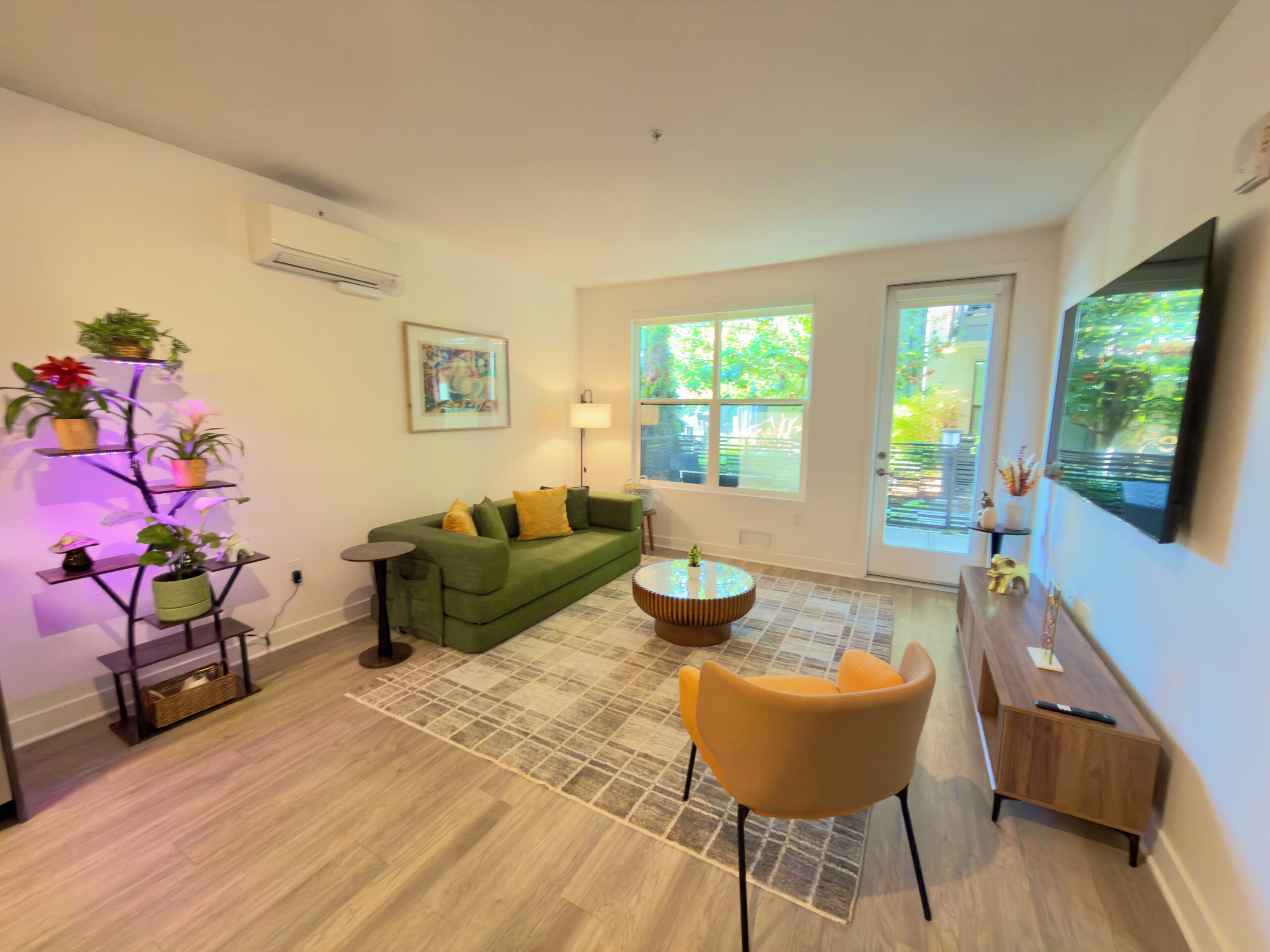 A living room with a green sofa, a small round table, a yellow chair, a floor lamp, and a wall-mounted TV.