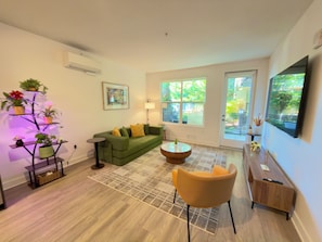 A living room with a green sofa, a small round table, a yellow chair, a floor lamp, and a wall-mounted TV. - Modern, 1-bedroom, near Stanford and Meta, with parking. (Menlo Park)
