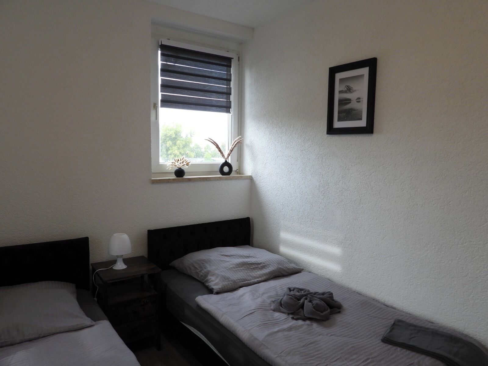 Ein kleines Zimmer mit einem Einzelbett, einem Fenster, einem Nachttisch mit Lampe und einem gerahmten Bild an der Wand.