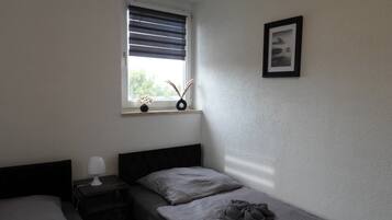 Ein kleines Zimmer mit einem Einzelbett, einem Fenster, einem Nachttisch mit Lampe und einem gerahmten Bild an der Wand.