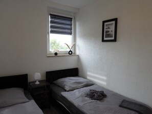 Una stanza piccola con un letto singolo, una finestra, un comodino con una lampada e un quadro appeso al muro.