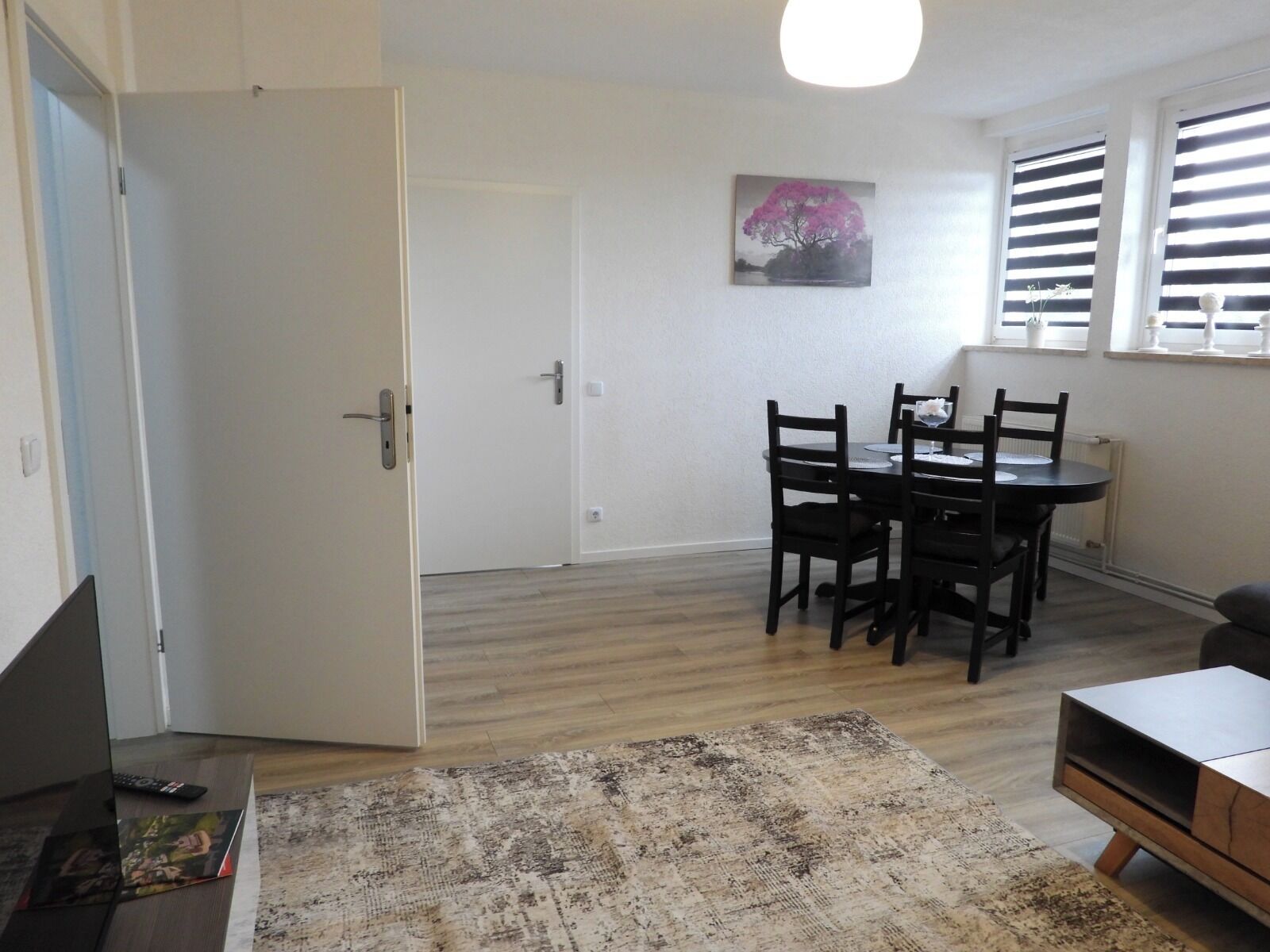 Ein Zimmer mit Esstisch, Stühlen, Teppich, Fernseher und einer Tür zu einem weiteren Raum.