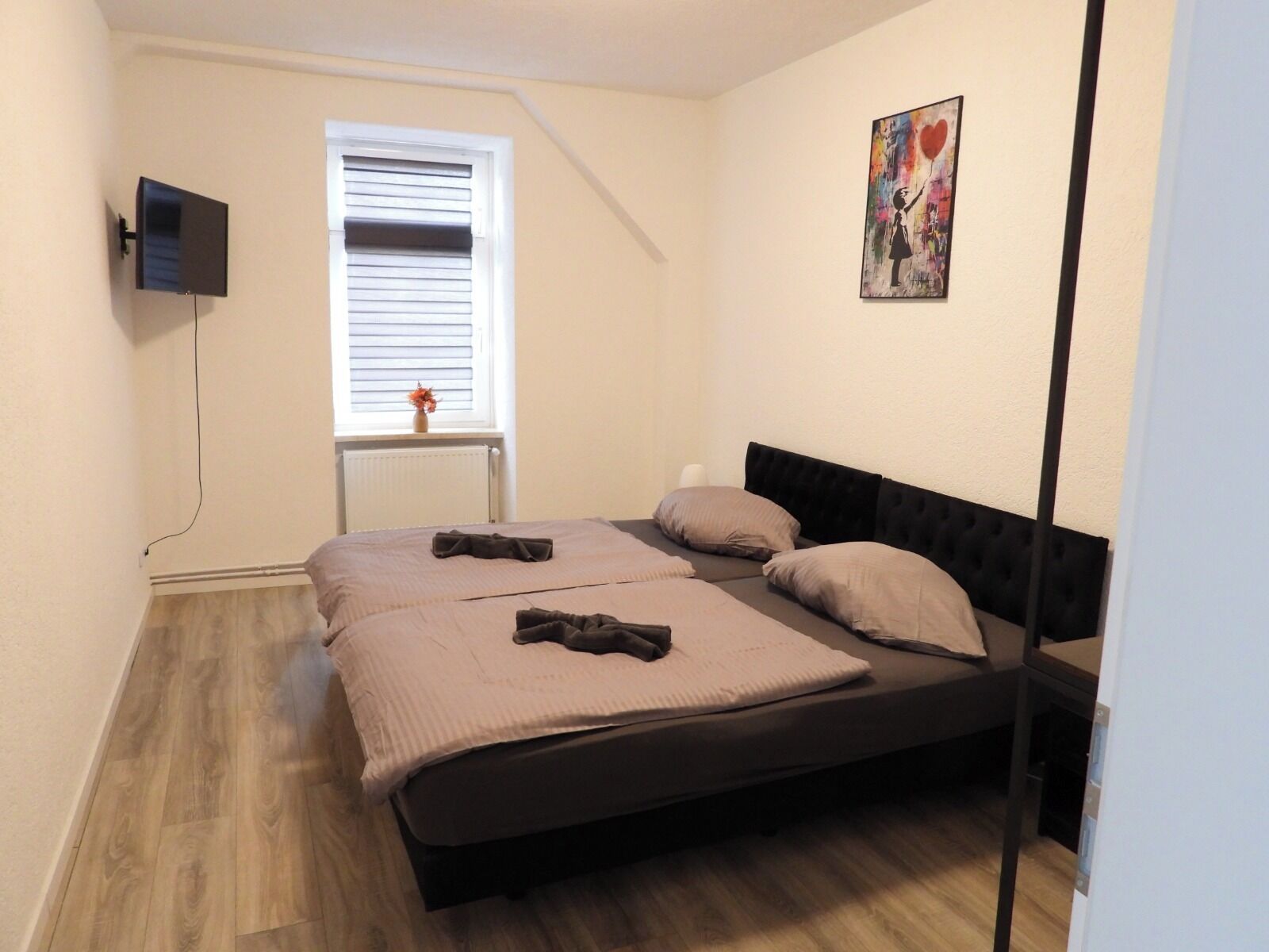 Ein Schlafzimmer mit einem Bett, einem Fenster mit Jalousien, einem Wand-Fernseher und einem Gemälde.