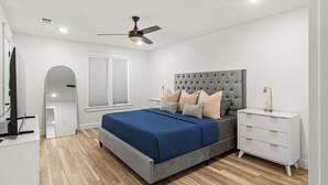 Une chambre à coucher comprenant un lit, des tables de chevet, une commode, un ventilateur de plafond et une fenêtre avec des stores.