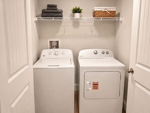 Une buanderie équipée de deux machines à laver et de deux sèche-linge à chargement frontal, tous deux de couleur blanche, d’une étagère où sont pliées des serviettes, d’une plante et d’un panier.