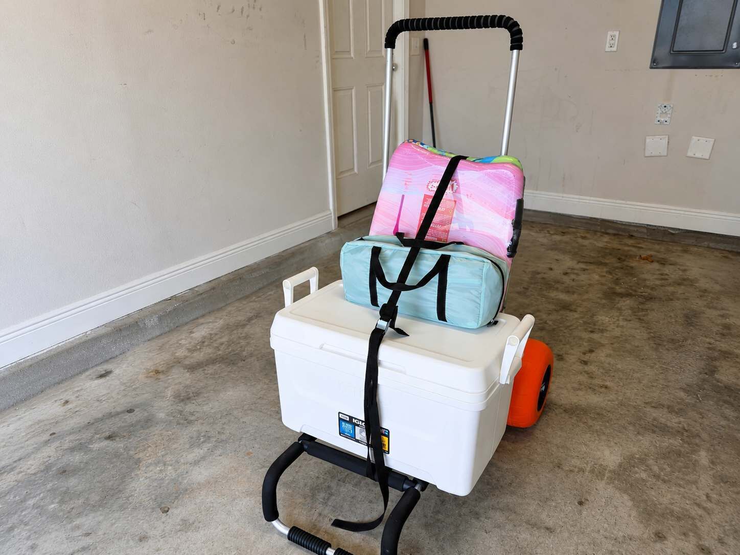Un carrito de equipaje con base blanca y un asa, que contiene una bolsa azul y una rosa, en una habitación con piso de concreto y una puerta al fondo.