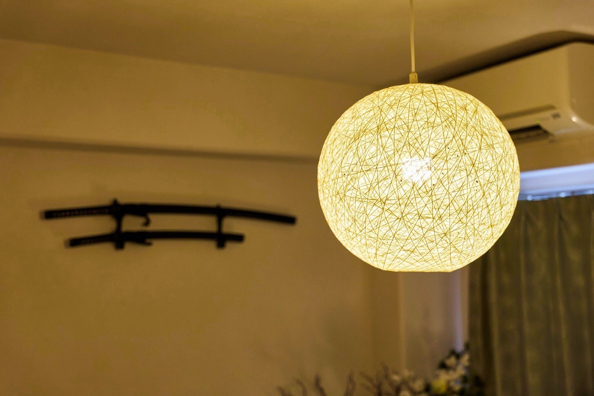 A spherical light fixture with a geometric pattern, hanging from a ceiling in a room with a plain wall and a visible air conditioning unit.