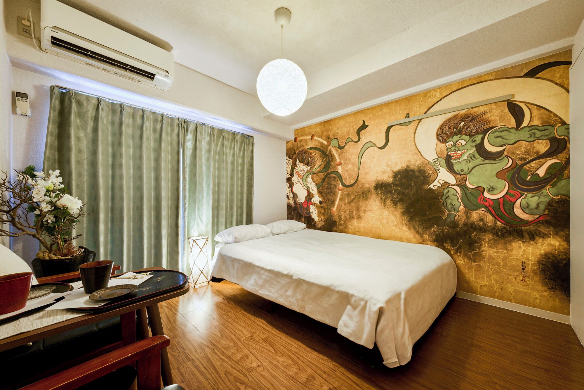 A hotel room with a bed, a wall mural, a desk with a vase of flowers, and an air conditioning unit.