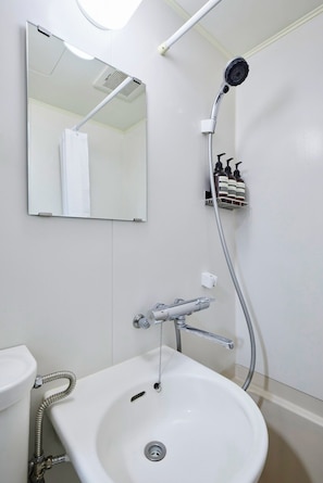 Un baño con lavamanos blanco, un espejo, un cabezal de ducha de mano y dispensadores de champú y acondicionador montados en la pared.