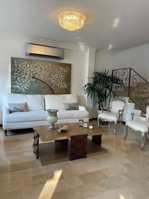 A living room with a white sofa, a coffee table, and two chairs. There is a large painting on the wall and a plant in the corner.