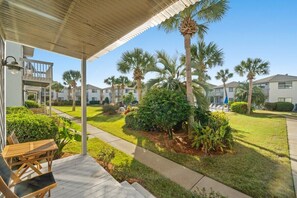 A covered outdoor area with a wooden deck, seating, and a pathway leading to a residential building complex with palm trees and well-maintained lawns. - Modern Retreat - Pool  - Walk to Beach (Miramar Beach)
