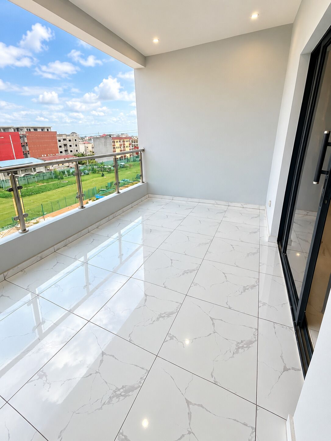 A balcony with a view of a green sports field and buildings, tiled floor, and a glass door.