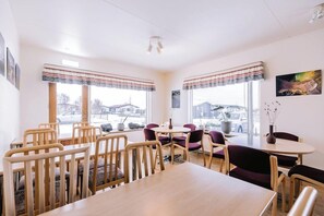 A dining area with wooden tables and chairs, large windows with curtains, and a view of the outside. - Hlíð Cottages (Myvatn)