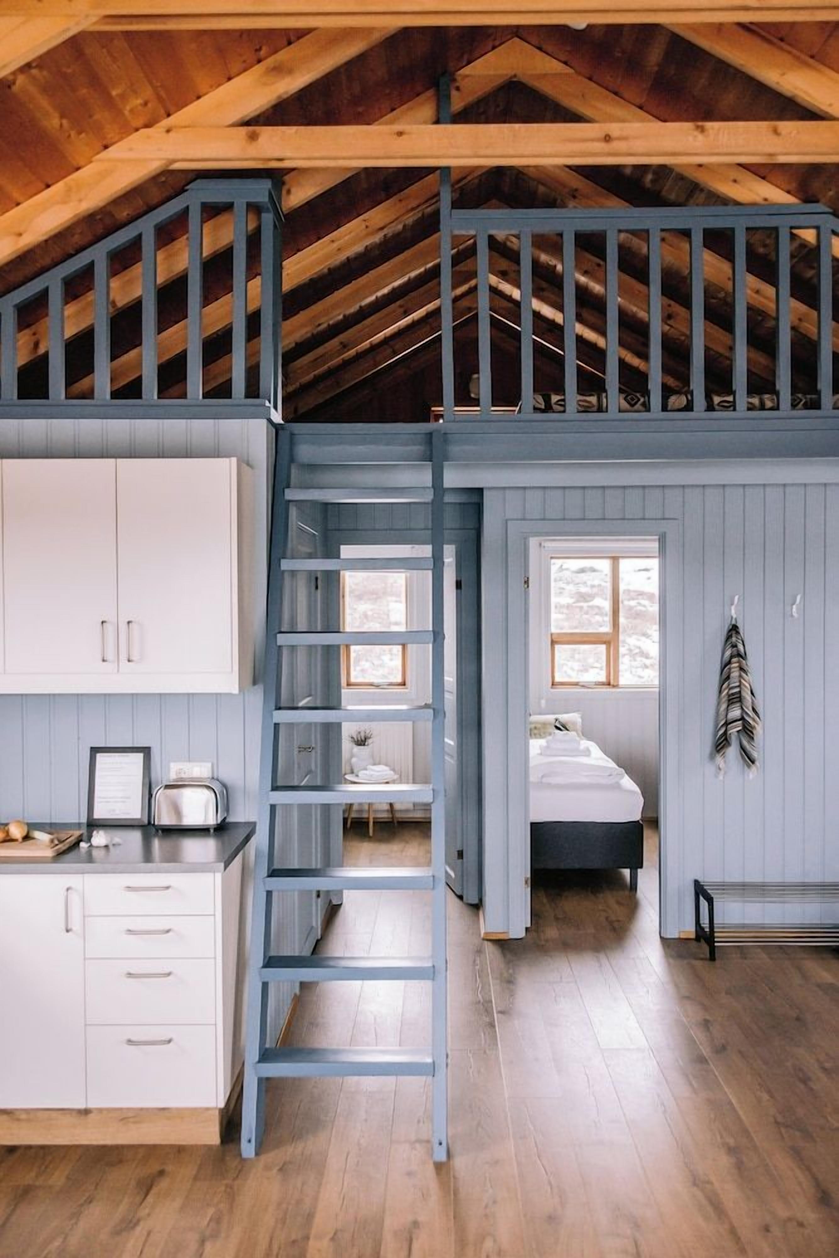Quarto estilo loft com teto de madeira, cama no mezanino, escada, janela e mesa de cabeceira.