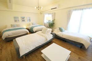 Quarto de hotel com três camas de solteiro, uma mesa de centro branca e uma janela grande com cortinas.