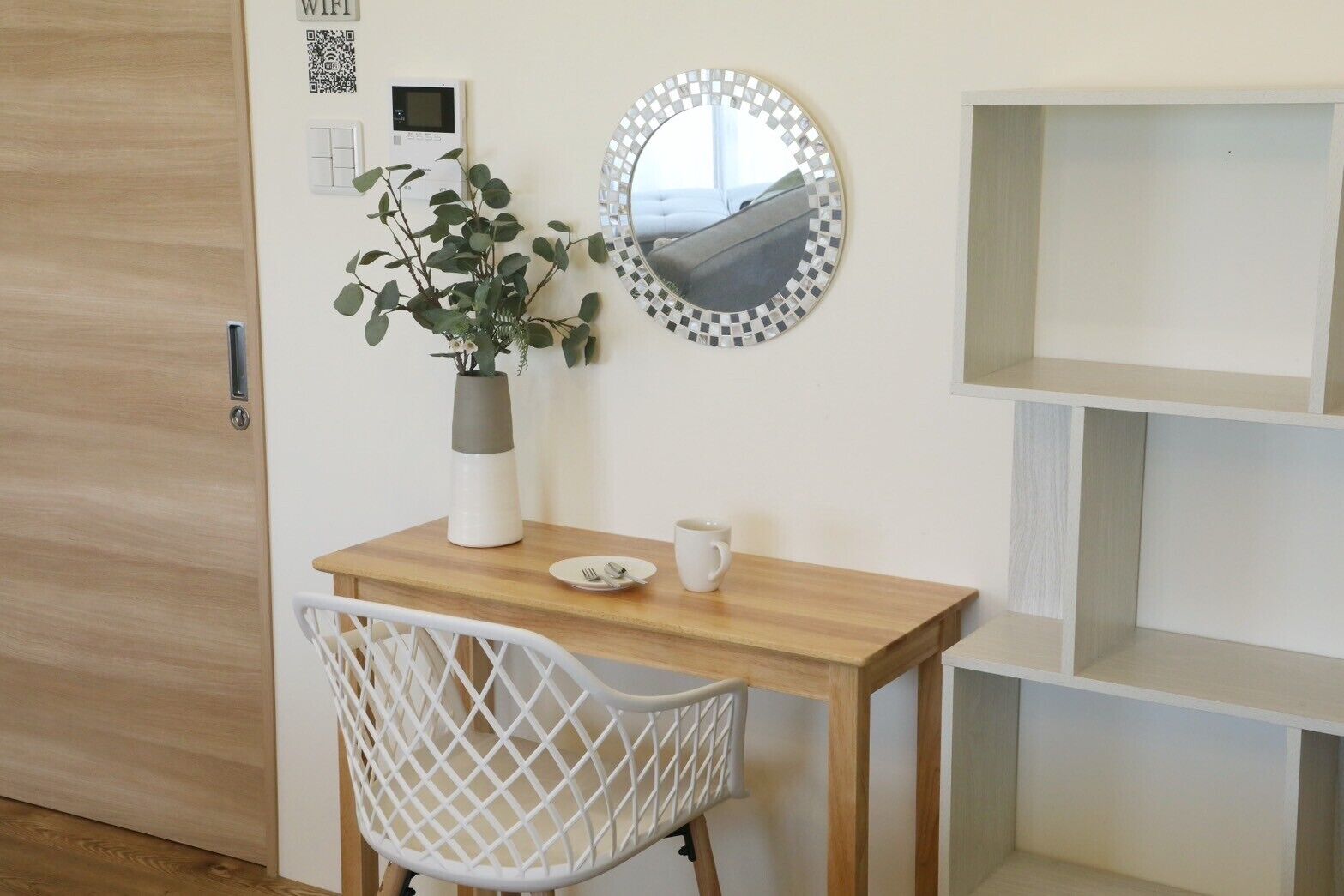 A room with a wooden desk, a white chair, a mirror, a vase with a plant, and a QR code on the wall.