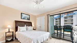 Quarto com cama, luminárias de cabeceira, ventilador de teto, porta que dá para uma varanda e quadro na parede.