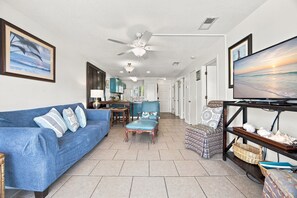A living room with a sofa, a television, a ceiling fan, and a view of the ocean through the window. - Romar Beach 108 Blue Tides (Orange Beach)