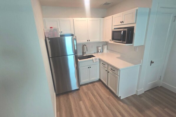 Una cocina compacta con muebles blancos, un refrigerador de acero inoxidable, un microondas y una cafetera.