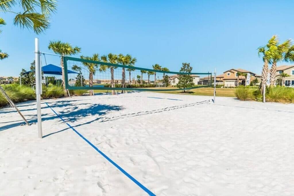 A beach volleyball court with a net, palm trees, and houses in the background.