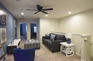 A living room with a dark grey sofa, a blue armchair, a plaid ottoman, a white side table with a lamp, and a ceiling fan. - Princess/SuperHero's 5-bedroom home near Disney (Kissimmee)