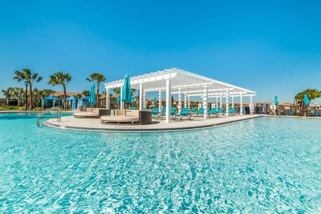 A clear blue pool with a white pergola and lounge chairs, surrounded by palm trees and umbrellas.