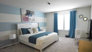 Une chambre à coucher comprenant un lit, une tête de lit, une fenêtre avec des rideaux, un berceau et un téléviseur fixé au mur.
