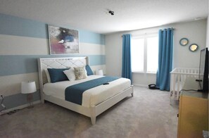 Un dormitorio con cama, cabecera, ventana con cortinas, cuna y televisor montado en la pared.
