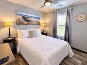 Ein Hotelzimmer mit einem Bett, zwei Nachttischlampen, einem Bild von einem Boot am Strand, einer Wanduhr und einem Fenster mit Vorhängen.