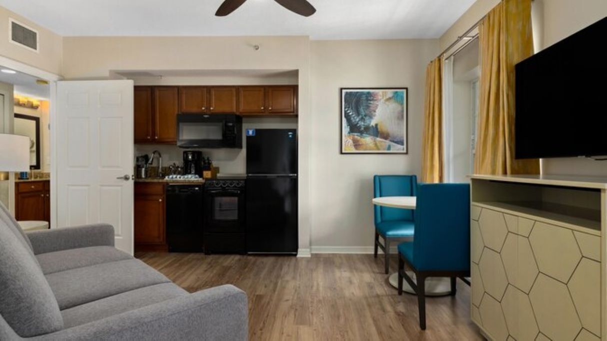 A compact living space with a kitchenette, a dining area with blue chairs, and a flat-screen TV mounted on the wall.
