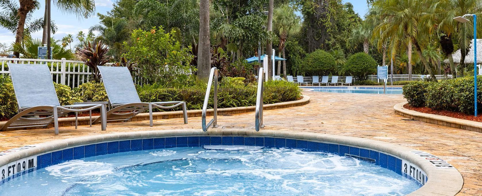 A hot tub surrounded by lounge chairs and a poolside area with a white fence and tropical plants.