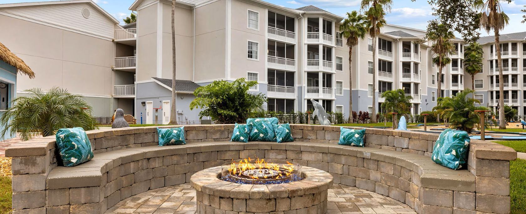 A circular stone fire pit with seating, surrounded by a stone wall, in a courtyard with a pool and palm trees.