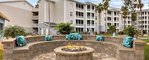 A circular stone fire pit with seating, surrounded by a stone wall, in a courtyard with a pool and palm trees. - Cozy, Fun & Exciting resort to stay at ! (Kissimmee)