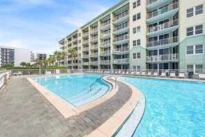 A swimming pool with a curved edge, surrounded by a paved walkway, and adjacent to a multi-story building with balconies. - Gulf View Condo with Shared Pool & Beach Access (Fort Walton Beach)