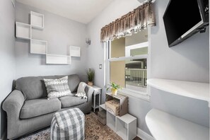 A modern living room with a grey sofa, a small side table, a potted plant, and a window with a view of the outside. - Gulf View Condo with Shared Pool & Beach Access (Fort Walton Beach)