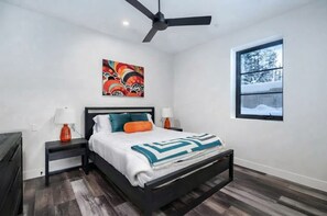 Ein Schlafzimmer mit Bett, Deckenventilator, Fenster mit Blick auf Bäume und einem Bild an der Wand.