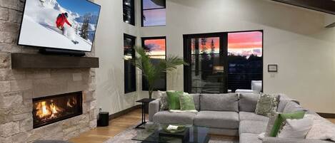 Uma sala de estar moderna com lareira, uma TV de tela grande e um sofá modular cinza, além de uma mesa de centro.