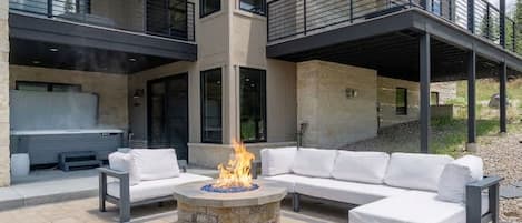 Un espacio exterior moderno con una chimenea de piedra, muebles de jardín blancos y un edificio de dos plantas con balcones.