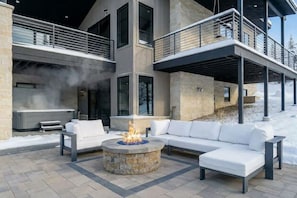 Ein moderner Außenbereich mit Feuerstelle aus Stein, weißer Gartenmöbel und Blick auf ein mehrgeschossiges Gebäude.