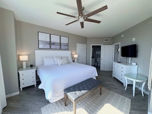 A bedroom with a bed, bedside tables, a bench, a ceiling fan, a TV, and a bathroom visible through an open door. - Coastal Condo with Shared Pools! (Destin)