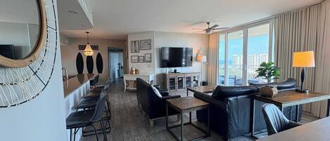 A modern hotel room with a dining area, a living room with a sofa and a TV, and a balcony with curtains.