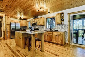 A kitchen with wooden cabinets, a stainless steel refrigerator, a granite countertop, and a large island with stools. - New! Mtn Views with Hot Tub and Fireplace, Fire Pit, Game Room & Work Desk. (Ellijay)