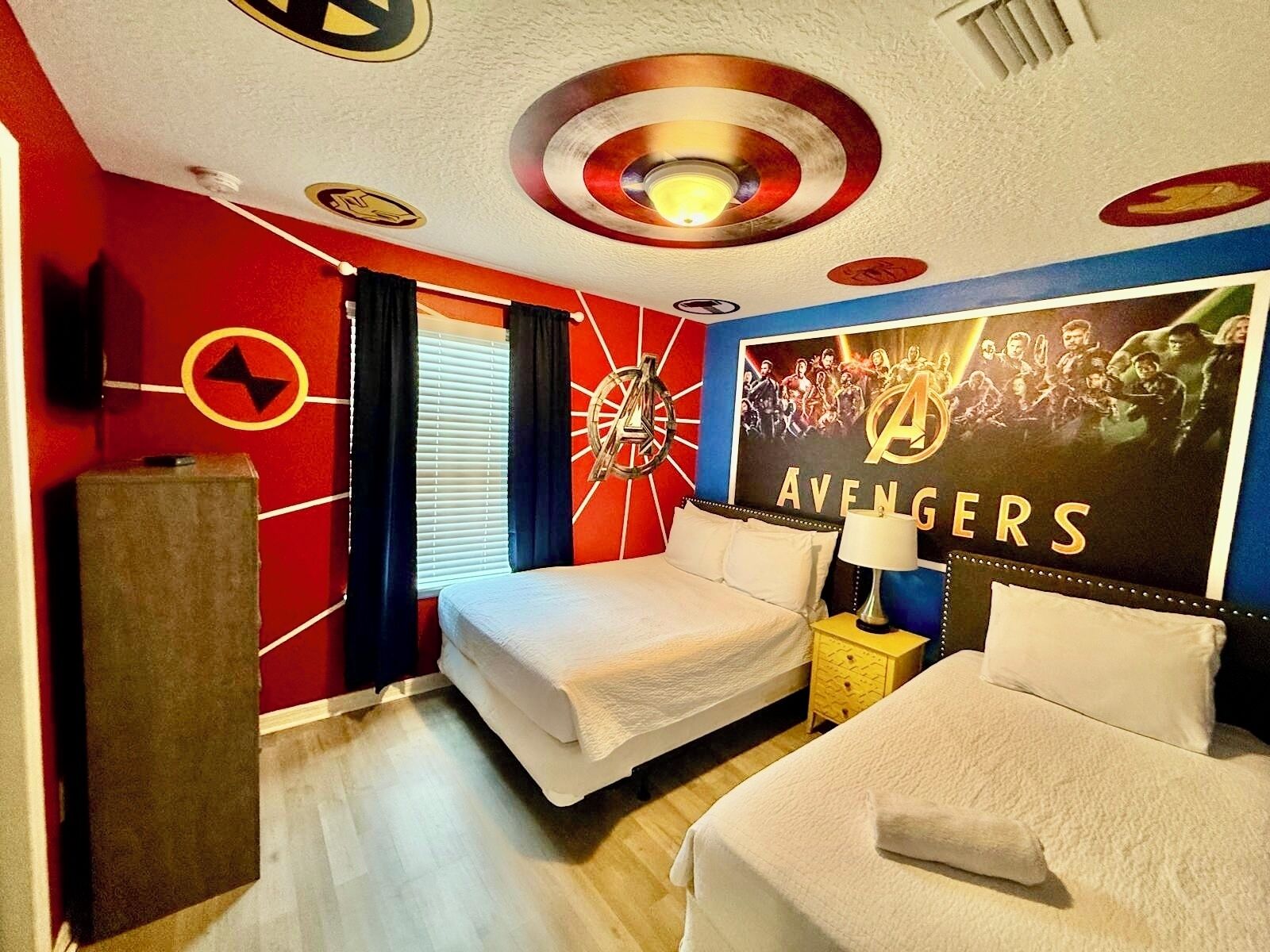 A hotel room with two beds, a large Avengers poster on the wall, and a unique ceiling design.
