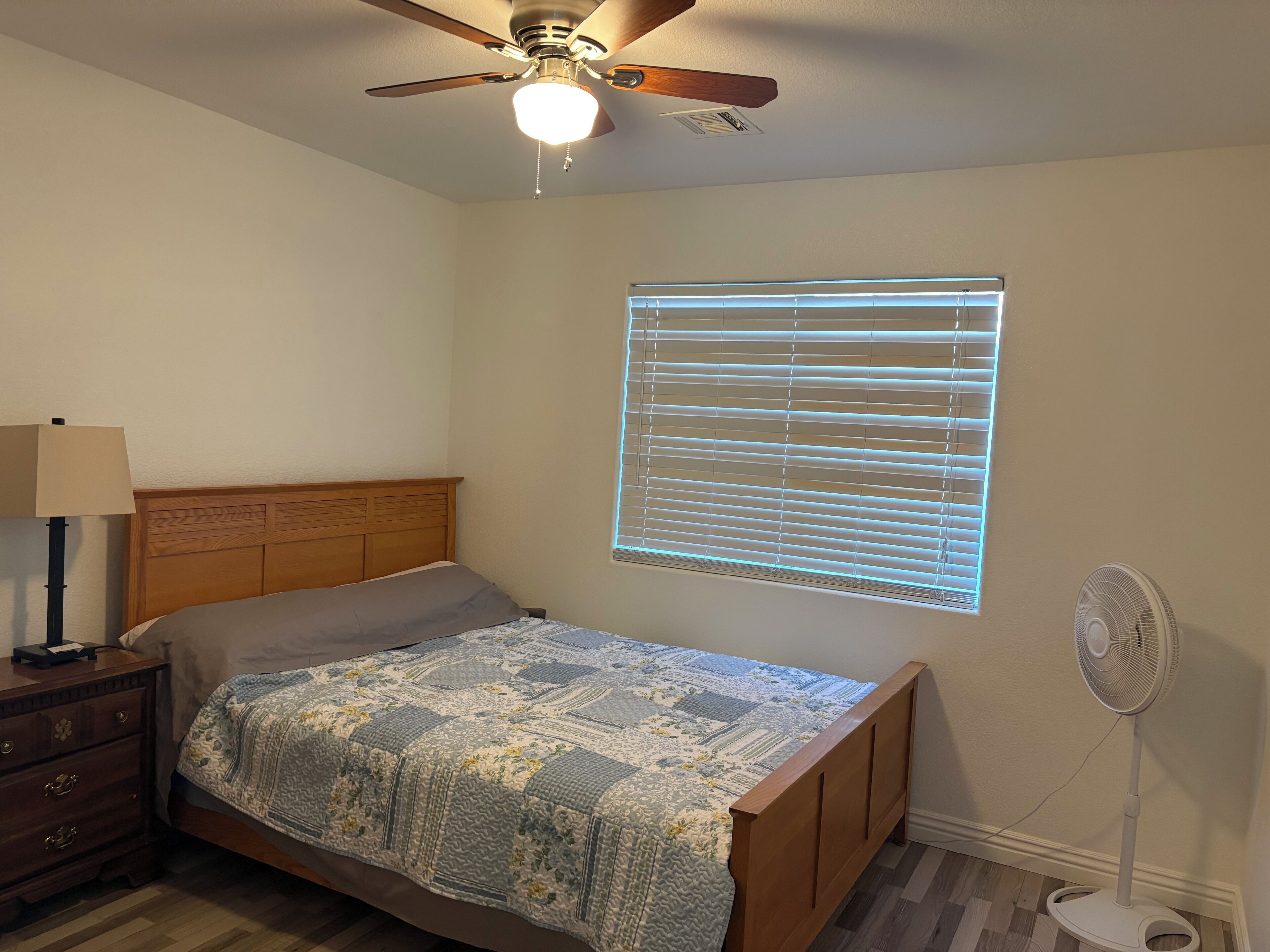 Ein Schlafzimmer mit einem Bett, einem Nachttisch, einer Lampe, einem Deckenventilator, einem Fenster mit Jalousien und einem Standventilator.